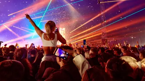 Pavilion Festival A view from the crowd at a music festival, with a dramatic lights show being displayed. Some people are filming it on their phones, while a woman is sitting on someone's shoulders with her arms in the air.