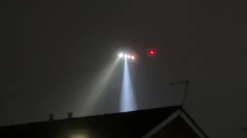 A picture of a helicopter above the rooftops. It is very dark and two wide white beams shine down behind the roofs. There are four other lights on the helicopter and two red ones.