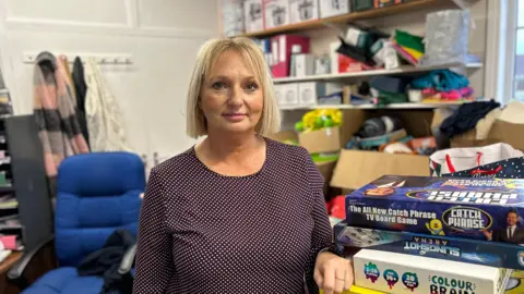 Jo Ramsay, who has a blonde bob haircut and is wearing a dark blue and white spotted dress, standing in her office with shelving and a blue office swivel chair behind her