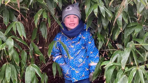 Evan standing under the cover of a large bush smiling at the camera. He is wearing blue jeans, a shiny blue and yellow jacket, a blue scarf and a grey bobble hat