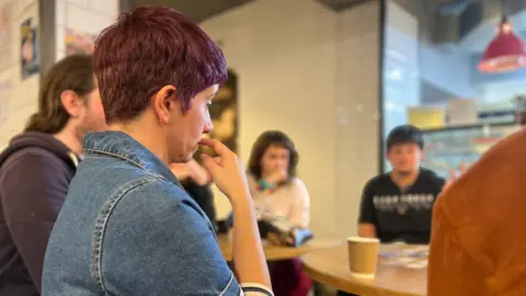 A group of people seated around a wooden table in a casual café setting. The person in the foreground is wearing a denim dress with striped sleeves and has short, dark reddish hair. A paper coffee cup is on the table, and the background shows white tiled walls and a hanging red light fixture.