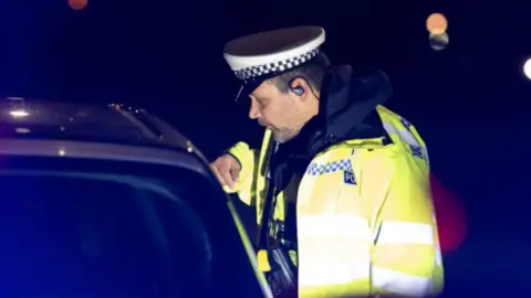 A police officer pictured speaking to the driver of a vehicle which has been stopped. The officer is leaning his right hand on the car and is wearing a high-vis jacket with police cap.