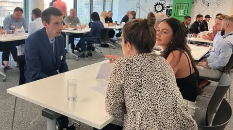 Danny sitting at a table being interviewed by two women - there are a lot of other people at tables doing the same in the background