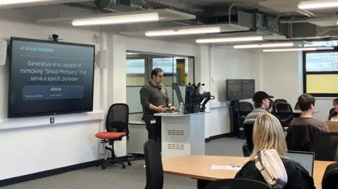 Lecturer Dr Ben Siu stood behind a tall desk leading the class on AI, in front of a large screen.