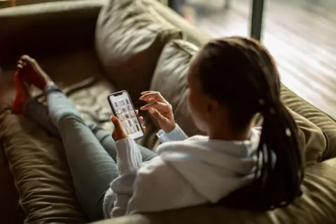 A woman with long dark hair pulled back from her face lies on a couch shopping on her phone