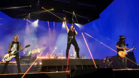 Getty Images Three members of a rock band on a stage. Red strobe lighting is lighting up the stage. The lead singer, a man in black trousers and a black waistcoat is jumping up in the air and two guitarists are standing on either side of him.