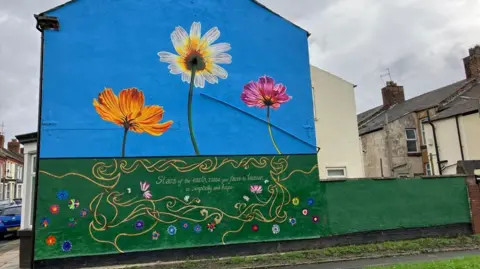 BBC A mural features blue sky and green grass, with numerous colourful flowers and three larger flowers against the backdrop of the sky. The words 'Stars of the earth, raise your faces to heaven in simplicity and hope' are included in the green section.