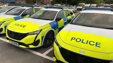 A line of three Wiltshire Police Peugeot estates, with blue lights on top, and fluorescent stickers around the car for visibility. The word POLICE is printed on each bonnet.