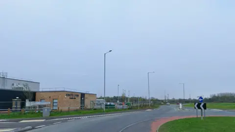 Borough Council of King's Lynn and West Norfolk A road leads up to a roundabout with grass growing in the middle and a red ring around it. Grey buildings can be seen in the distance.