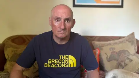 A man with a bald head and a navy T-shirt with yellow writing on it is looking at the camera. He is sitting on a beige sofa with cusions. The wall behind him is magnolia and the base of a framed picture can be seen in the top right corner.