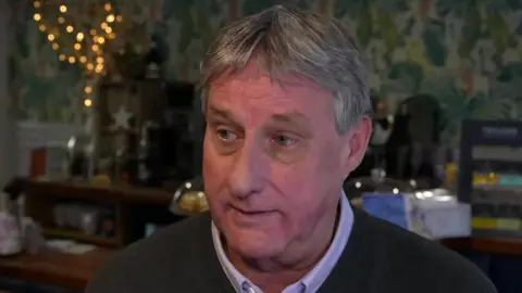 Jim, who is clean shaven with short grey hair, sits in front of a cafe counter and colourful wallpaer. 