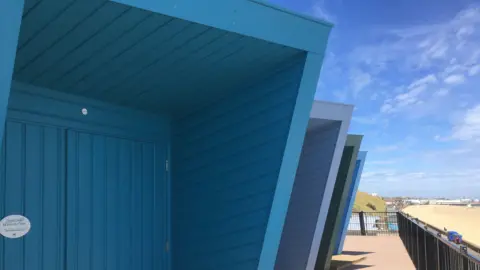 GUY CAMPBELL/BBC Modern beach huts painted in different shades of blue are facing towards the sandy seafront  