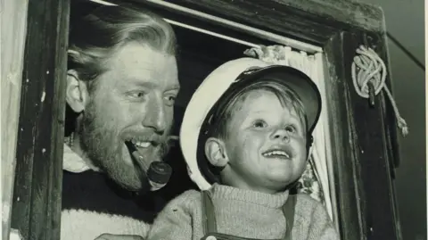 Paul Ainsworth A black and white picture of a young Paul Ainsworth wearing a sailor's cap leaning out the window of a boat with his father Mark stood behind him. Mark is wearing a wool, knitted humper with a smoking pipe in his mouth, smiling and glancing at Paul. 