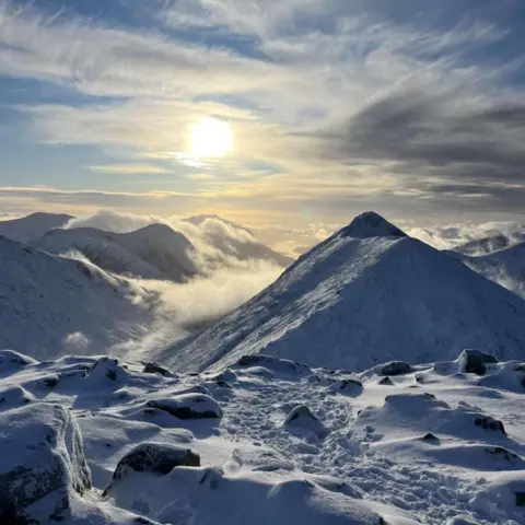 Lachlan Moffett The sun shines behind clouds above a snowy mountain landscape. Low cloud hangs in glens between the mountains.