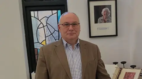 Head and torso shot of a middle-aged man with a receding hairline and serious expression. He is wearing a brown jacked and blue and white checked shirt. Behind him to the right a portrait of a woman is on the wall and there is an open book on a stand next to his elbow.