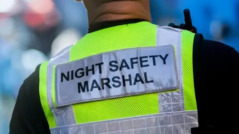 Sussex Police A close up of a male marshal's back wearing a black top and high vis jacket with a night safety marshal sign on the back