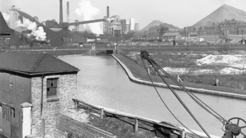 Staffordshire Record Office A black and white photograph showing a body of water with an industrial site in the background. There are plumes of smoke or vapour emanating from the buildings. The brick building in the foreground has broken windows on its upper floor. There is also a crane in the foreground.