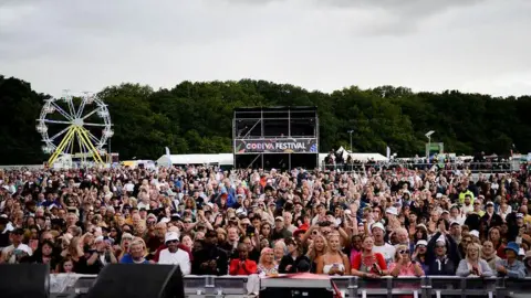 A crowd of hundreds of people stand at the front of the main stage at the park. A ferris wheel can be seen behind then, as well as a stand with the words Godiva Festival.