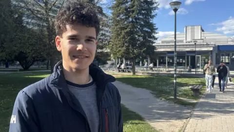 A young man with dark curly hair stands in front of a lawn and a modern building