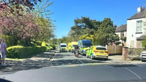 Ambulances and police vans line a residential road with semi-detached houses on the right and hedgerows on the left. 