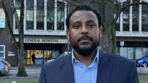Doctor Awilly Chofle smiling at the camera. He is stood in front of Jersey General Hospital and is wearing a blue suit jacket with a light blue shirt. 