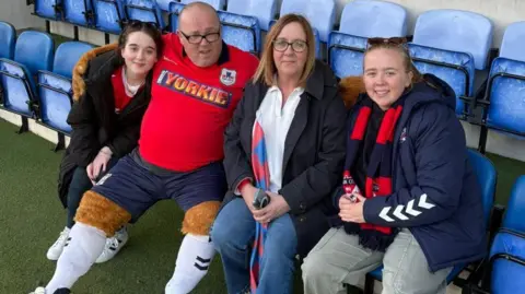 Supplied A family of two young women, mum and dad, sitting on fold-down chairs in a football stadium. The Dad is wearing a large lion mascot costume with "Yorkie" written on the shirt.