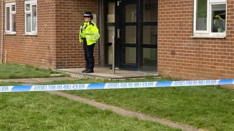 SimonThake/BBC A female police officer wearing a yellow jacket over her black uniform and hat stands in front of the entrance to a block of flats