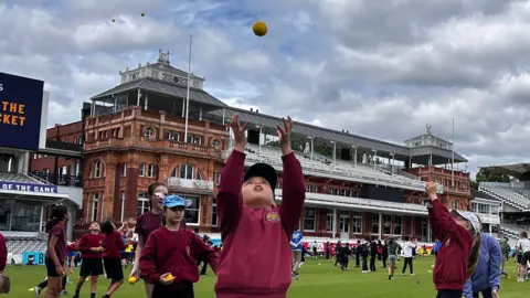 A group of children wearing caps and burgundy sweatshirts and standing on the pitch in Lords cricket grounds, throwing balls in the air.