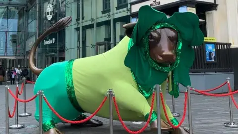 The bullring bulll dressed up in a green outfit with a shamrock headdress, behind red rope railings.