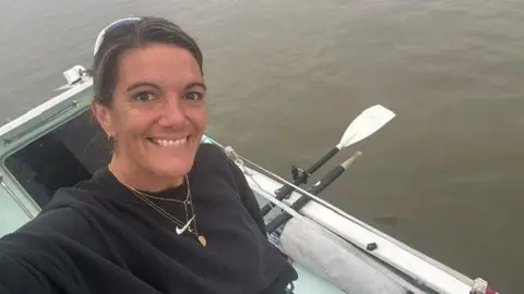Clare O'Reilley A woman smiling for a selfie with her arm above her head to take a looking down photo on a rowing boat with seas below her.