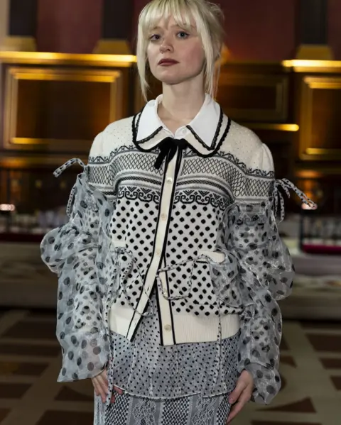 Jordan Pettitt/PA Wire Maisie Peters, a young women with white-blonde hair, wears a black and white blouse and skirt with lots of clashing patterns. She is stood in a decorated hall