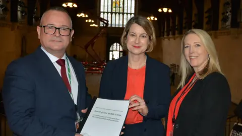 Lee Barron on the left in a suit and tie handing a report to Helen Hayes, who in a red top and blue jacket. Kay Sammon is on her right in a black jacket and red scarf. All are in Parliament and looking at the camera.