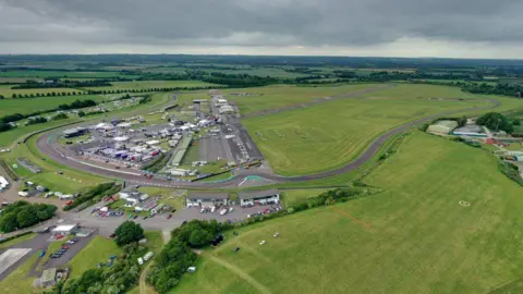 Aerial view of Thruxton Aerodrome and motor racing circuit on a cloudy day. There are two asphalt runways in a cross shape in the middle of a large area of grass. On the left of the picture are the hangars and circuit buildings. Lines of small aircraft are parked on the grass and on the tarmac taxiway. The race track runs around the edge of the site.