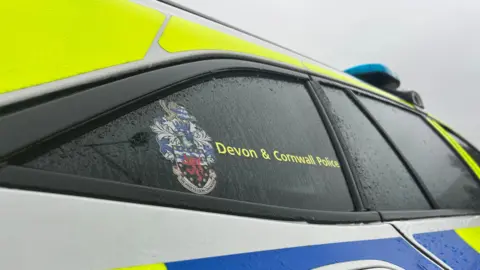 Exterior of a Devon and Cornwall Police car. The police logo is displayed on the car's window. Rain droplets are running down. 