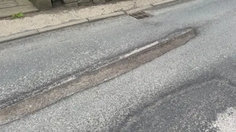 Matthew Lynch A pothole in the shape of a long line and next to another pothole of a circular shape on the same road.