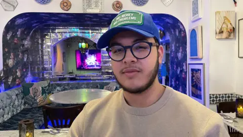 Man with glasses and beard wearing a blue and green cap saying 'no money no problems' in white letters