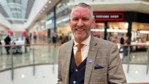 Head and shoulders photo of a man with short gray hair and stubble, wearing a brown blazer, yellow tie and blue cardigan, smiling at the camera. It is in a shopping center with stores behind it.