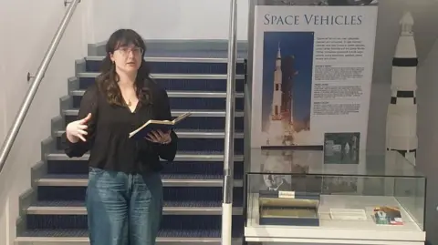 Armagh Planetarium A woman standing speaking with a notebook open in one hand while the other gestures. She has dark hair and is wearing glasses, a black top and jeans. She is standing in front of stairs beside a museum display.