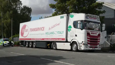 The photo shows a predominantly white articulated lorry, which is stationary. It has red lettering on the side with the name of the haulage company, Cattafi Transervice, and green lettering which says International transport & logistics Holland Italy. Behind the lorry, there is a police car and a police officer. 