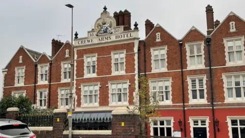 An image of Crewe Arms Hotel - a large 19th Century red brick building with a large crest at the top which says Crewe Arms Hotel.