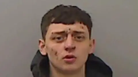 Mugshot of Harbron. He is clean shaven with short dark hair. He has cuts all over his face and appears to have bruises under each eye.