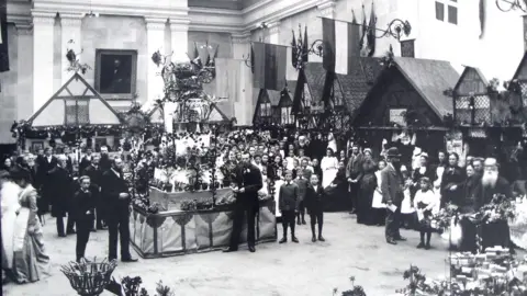 The Corn Hall Diss A black and white photo from the Victorian era shows several dozen men, women and children inside Diss Corn Hall, standing around a central display of what look like pot plants. Around the periphery are several timbered replicas of medieval German-style cottages.