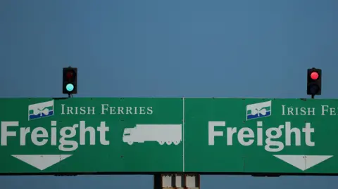 Reuters Two green signs. Both signs have the words 'Irish Ferries Freight' written on them. Above each sign is a traffic light. The traffic light on the left is green, while the traffic light on the right is red. 