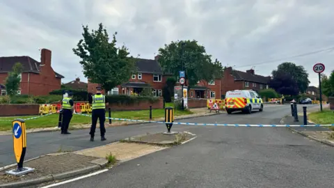 BBC/Emily Johnson Two police officers stand in front of a barrier at the top of Bellfarm Avenue