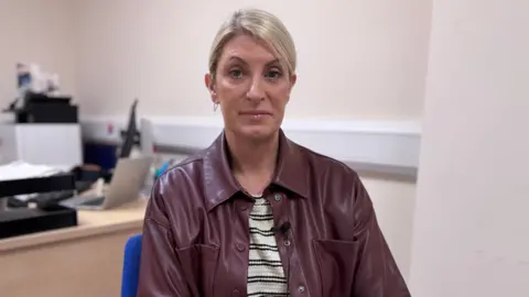 Linda has blond hair to her shoulders.
She is wearing a burgundy leather coat and a cream and black striped shirt.
She is sat in an office with a desk and computer visible in the background