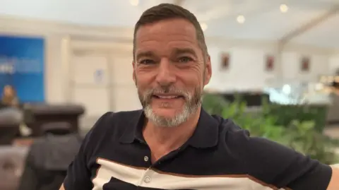 A close-up of Fred Sirieix, who has a greying beard and is smiling into camera wearing a black polo shirt with a cream stripe across the chest