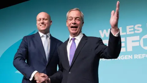 Getty Images Dan Thomas and Nigel Farage standing front of a Reform UK logo. Farage has one hand in the air waving to people.