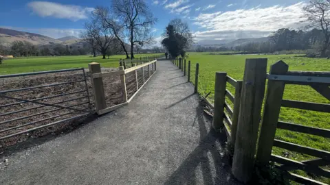 LDNPA A path stretches out between two fields with a metal gate on one side, a wooden one on the other and fencing either side. A few trees are in the distance and the hills of the Lake District rise up into a blue sky 