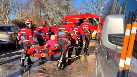 BCP Council Rescuers in red jackets transport a woman on a red stretcher to safety. She is sitting up and has blonde hair and a pink top on. 