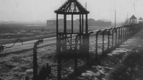 PATHE A black and white photograph of razor wire and guard towers at Auschwitz concentration camp